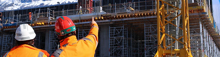 Two workers in front of a building site wearing protective gear.
